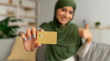 Happy young Arab woman holding credit card and showing thumb up gesture at home, selective focus