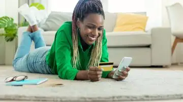 Excited black woman laying on floor carpet with cell phone and plastic credit card, ordering food or shopping online